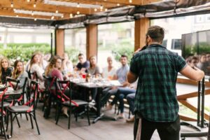 Man in plaid shirt speaking in front of group of people outdoors with food and drinks on the table.